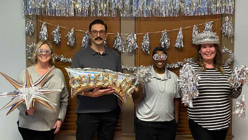 Express Employment Professionals team celebrates achieving the Platinum Circle of Excellence Award. The General Manager, Business Developer, and Employment Specialists hold metallic balloons and streamers with metallic decorations in the background. The team members also wear confetti metallic glasses and glitter hats.
