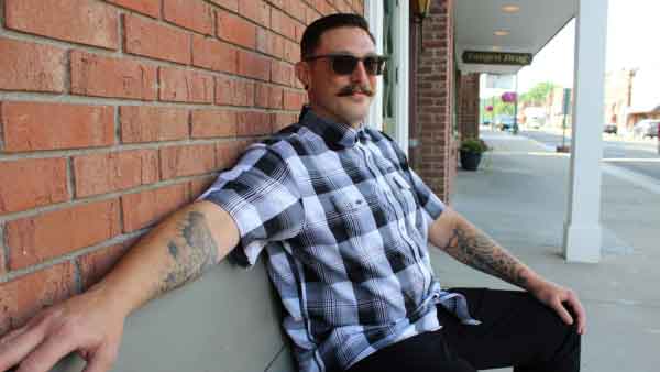 Business Developer wears button up shirt smiling and sitting on a bench against a brick wall with arm resting on back of bench in a downtown setting.