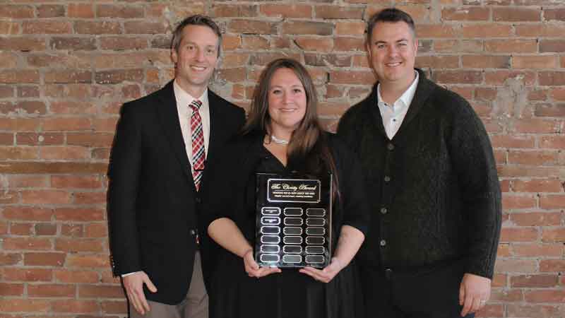 Office Manager stands between two Owners with gilded plaque award against a brick wall. All individuals are smiling and wearing professional attire. This is the Christy Rikli Legacy Award that is presented to one individual in the franchise group each year at the Kickoff event in January.