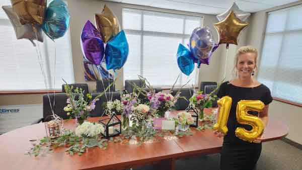 Owner celebrates 15 years with Express Employment Professionals. She is smiling and holding balloons in the shape of the number 15. There are bouquets of flowers and clusters of balloons on a table in the background.