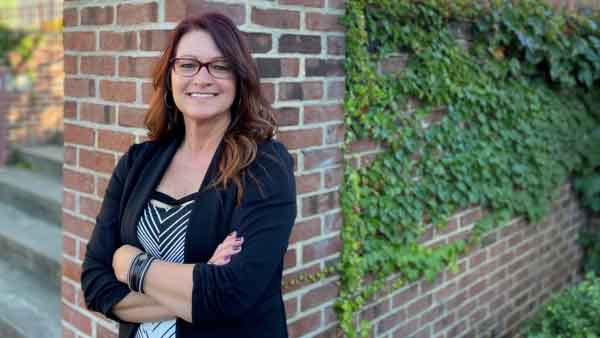 Front Office Coordinator / Payroll Specialist wearing professional attire leans against brick wall smiling with arms crossed.