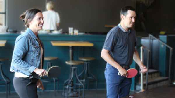 Specialized Recruiting Group Director of Account Management and Professional Recruiter wear casual attire and hold ping pong paddles at Punch Bowl Social as part of the Halftime event that takes place in July each year. The event space is filled with a ping pong table, foosball table, bowling alley, and more.