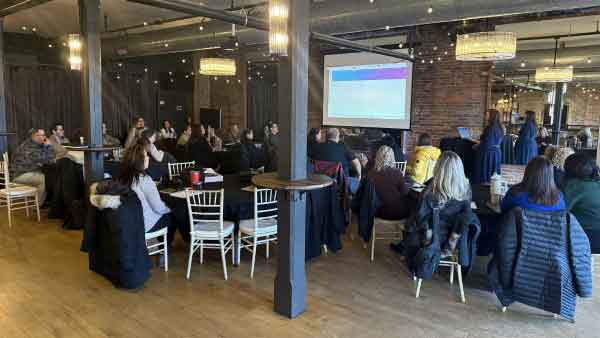 Franchise group of employees sit at round tables in an event space facing the front of the room during a presentation from the Marketing Manager. Wood floors, brick walls, and ambient lighting create a warm environment for the franchise group Kickoff event held in January each year.