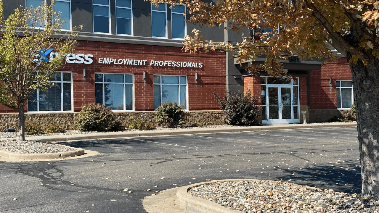 Photo of the exterior of the Express Employment Professionals Building in River Falls, WI. Includes the parking lot, a tree, and the red brick building with gray paint, white trim, and the Express Employment Professionals logo.