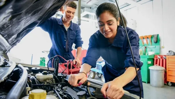 Two young adult mechanics (one man, one woman) in blue coveralls working together under a car hood in an auto shop.