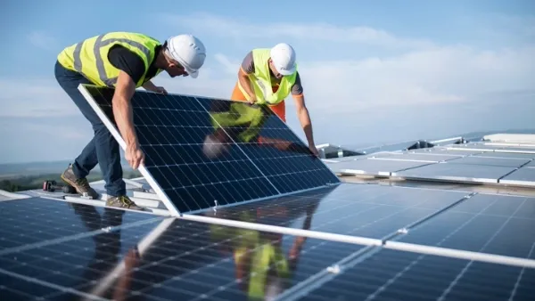 Two adult men in hard hats and safety vests installing solar panels on a rooftop under a clear sky.
