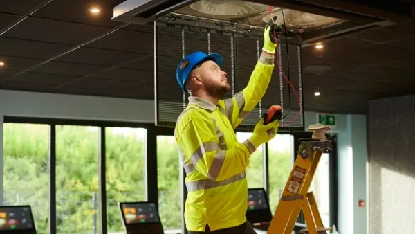 An adult male technician in a high-visibility jacket and blue hard hat standing on a ladder while repairing a ceiling unit in a gym with treadmills behind him.