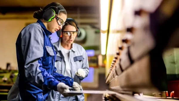 Two adult workers (appearing female) in protective eyewear, gloves, and earmuffs operating machinery in an industrial setting.