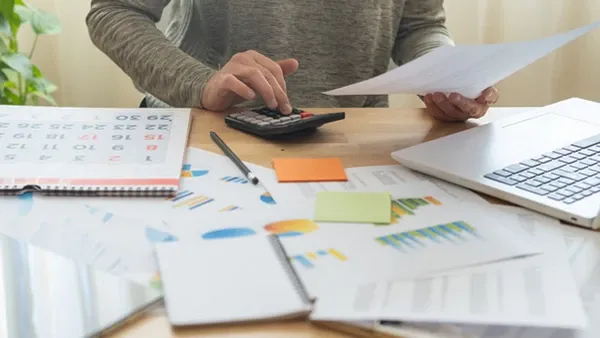 Photo of a person reviewing paperwork and using a calculator at a desk with charts, a laptop, a tablet, sticky notes, and a calendar.