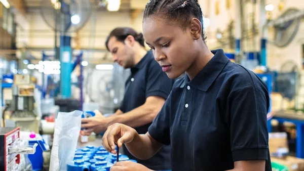 Photo of two assembly line workers in a factory setting; a foreground worker appearing female with hair in a braided bun wears a black short‑sleeve polo shirt and uses a hand tool to assemble blue mechanical components on a workbench, while a background worker appearing male in a black shirt performs a similar task along the same production line.