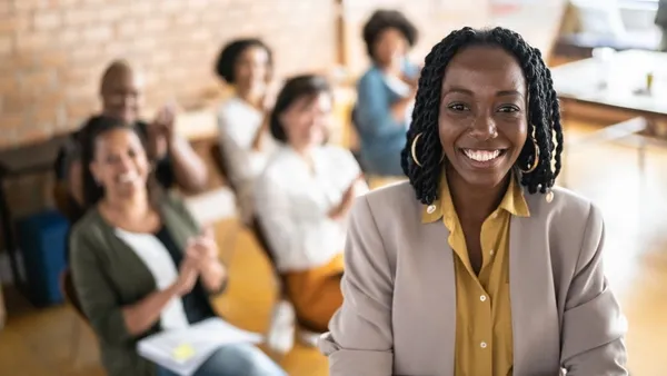 Photo of a Black woman standing in the foreground with arms crossed, wearing a light-colored blazer over a yellow shirt. In the background, several people are seated in a meeting, with a brick wall and sticky notes visible. A table with papers and a laptop is positioned near a window, suggesting a collaborative workspace.