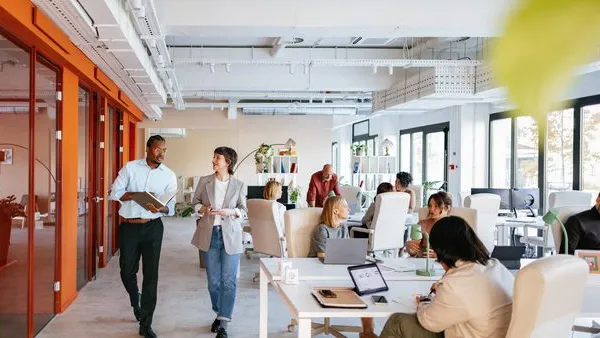 Photo of a modern open-plan office with several people working at desks and two individuals walking and talking in the foreground. The space features large windows, white walls, orange accents, and contemporary furniture.