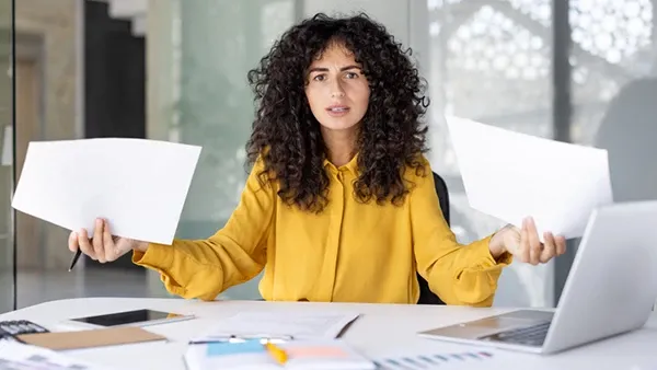 Photo of a person with curly dark hair wearing a yellow blouse, seated at an office desk holding two blank sheets of paper. A laptop, clipboard, calculator, and documents are spread across the desk, with a modern glass-walled office in the background.