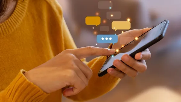 Photo of a woman with medium‑length dark hair wearing a mustard yellow sweater, using a smartphone indoors. She taps the screen with one hand while holding the phone in the other. Colorful illustrated chat bubbles float above the phone, symbolizing messaging or social interaction.