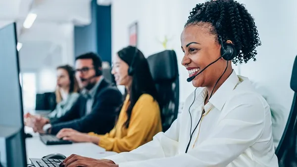 Photo of customer service representative wearing a white button-down shirt and headset, typing at a computer workstation in a modern call center, with three additional headset-wearing colleagues and monitors visible in the background.