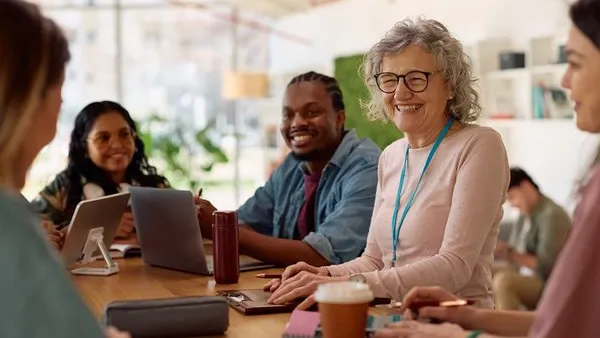 Group of diverse professionals engaged in a collaborative meeting around a table in a modern, well-lit office space. Participants are actively conversing and using laptops and tablets, with notebooks, coffee cups, and office supplies scattered across the table. Large windows provide natural light, enhancing the open and contemporary workspace.
