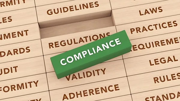 Photo of a green rectangular block labeled “COMPLIANCE” placed diagonally on stacked wooden blocks engraved with words including “REGULATIONS,” “GUIDELINES,” “LAWS,” “REQUIREMENTS,” “LEGAL,” “RULES,” “STANDARDS,” “VALIDITY,” “ADHERENCE,” “CONFORMITY,” and “AUDIT.”