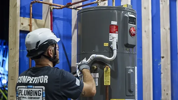 Photo of an adult man wearing a white safety helmet, gray gloves, and a black T‑shirt with the text “COMPETITOR” and “PLUMBING National Championship” on the back, installing a gray water heater connected to copper piping in an indoor construction setting.