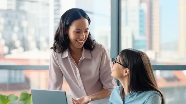 Two women collaborate at a modern office desk, with one standing and gesturing toward an open laptop while the other sits, listens, and learns. The workspace includes notebooks, a coffee cup, and a smartphone, and large windows behind them reveal a city skyline filled with natural light.