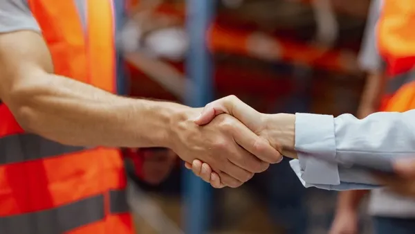 Photo of two people shaking hands in a warehouse. One man wears an orange safety vest and gray shirt; the other person wears a light blue long‑sleeve shirt. Shelving and another person in an orange vest are visible in the background.