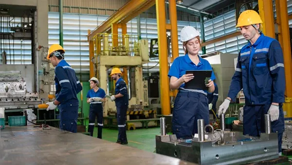 Photo of workers wearing blue uniforms and safety helmets operating machinery and reviewing information inside a manufacturing facility.