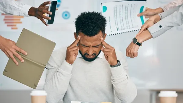Photo: Person in a light gray sweater seated at a desk, pressing fingers to temples while multiple coworkers’ hands extend a smartphone, folder, printed chart, and pointing gestures toward the person; office setting with documents, notebook, calculator, and coffee cups on the desk.