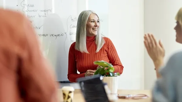 Photo of a woman with long gray hair wearing a red turtleneck sweater standing in front of a whiteboard, speaking to two people seated at a table. One person is clapping and another is partially visible in the foreground. Laptops, a mug, and a small plant are on the table.