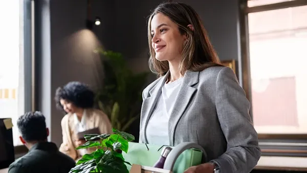 Photo of a person in a light gray blazer and white shirt carrying a box with a green plant and office supplies in a modern office; two other people are blurred in the background near desks and windows.