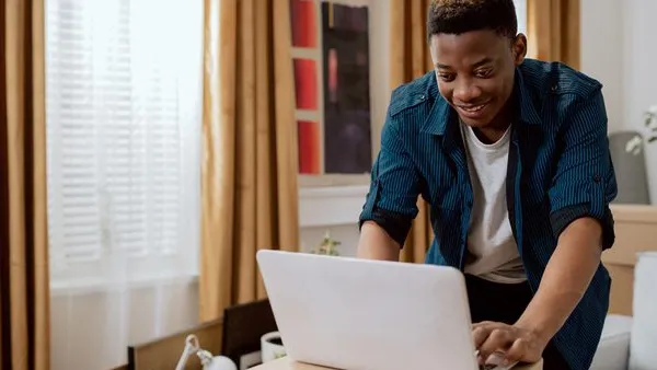 Black person working on a laptop placed on cardboard boxes in a room with golden curtains, surrounded by furniture, plants, and wall art.