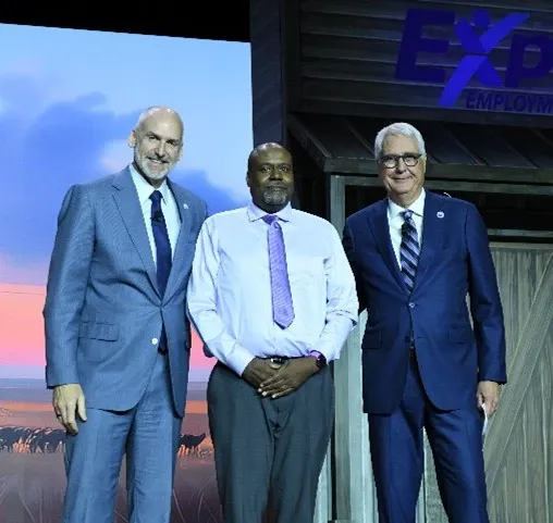 Photo of three adults standing on a stage in business attire. Person on the left wears a light blue suit and tie, person in the center wears a white dress shirt with a light purple tie and gray pants, and person on the right wears a dark blue suit with a striped tie and glasses. Background includes an illuminated stage backdrop and the partially visible logo text: “EXP Employment.” Provide your feedback on BizChat