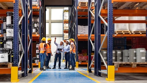 Photo of six adult workers—men and women—wearing yellow hard hats, orange safety vests, long pants, and work boots, standing in a warehouse aisle between tall pallet racks filled with boxed and bagged inventory; one person gestures upward while others look on, suggesting a team discussion or inspection.