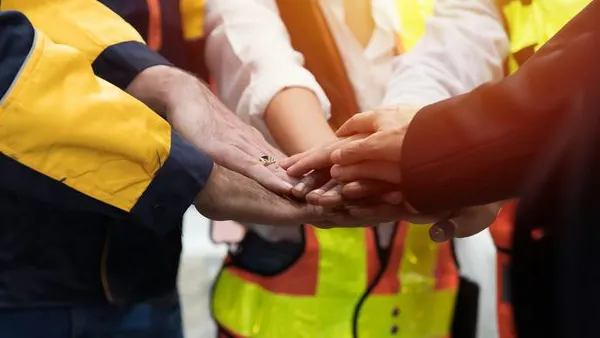 Group of workers in high-visibility safety vests and uniforms standing in a circle with hands stacked together, symbolizing teamwork, unity, and collaboration in an industrial or construction setting. The image reinforces themes of workplace solidarity, team performance, and leadership in hands-on environments.