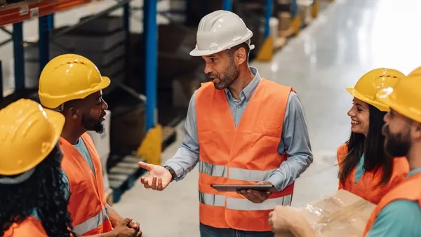 Photo of a group of workers in a warehouse wearing orange safety vests and yellow or white hard hats. A man in a white hard hat and orange vest stands at the center holding a tablet and speaking to the team. The others, including women and men, stand around him listening; one person on the right holds a cardboard box. Rows of shelves and inventory line the background.