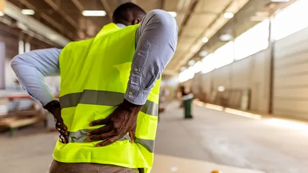 Photo of a person wearing a high-visibility yellow safety vest over a light blue long-sleeve shirt and brown pants, standing indoors in an industrial workspace with both hands placed on the lower back; a yellow hard hat and ear protection rest on a work surface in the foreground.