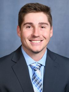 Professional photo of a young man wearing a navy suit jacket, a light blue shirt, and a tie with various shades of blue.