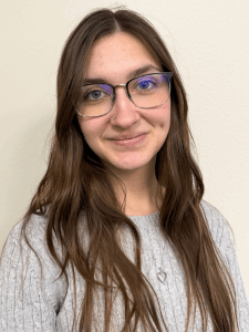 Professional photo of a young woman with long brown hair, glasses, and a light grey sweater